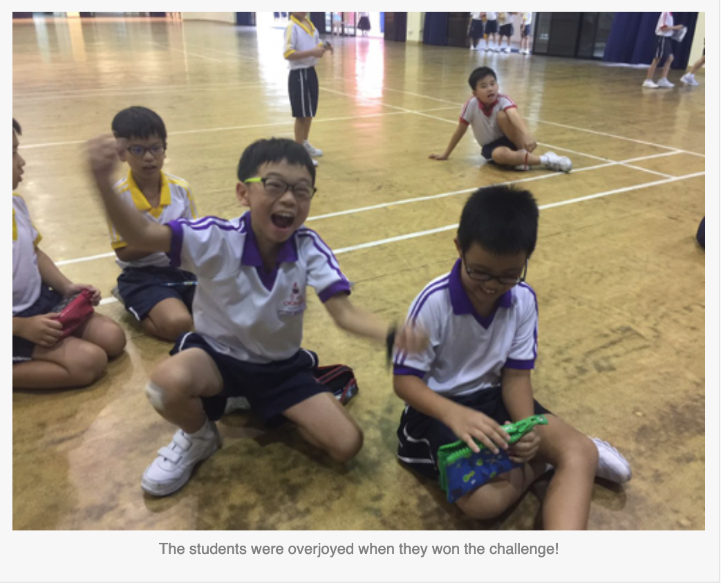Students in school uniforms sit on a gymnasium floor, one boy cheering with mouth open wide in excitement.